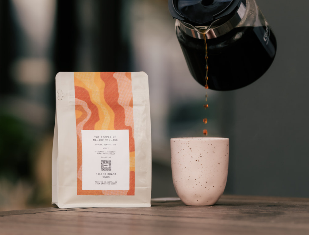 Person pouring coffee from a black coffee maker into a pink ceramic cup with a coffee bag in the foreground.